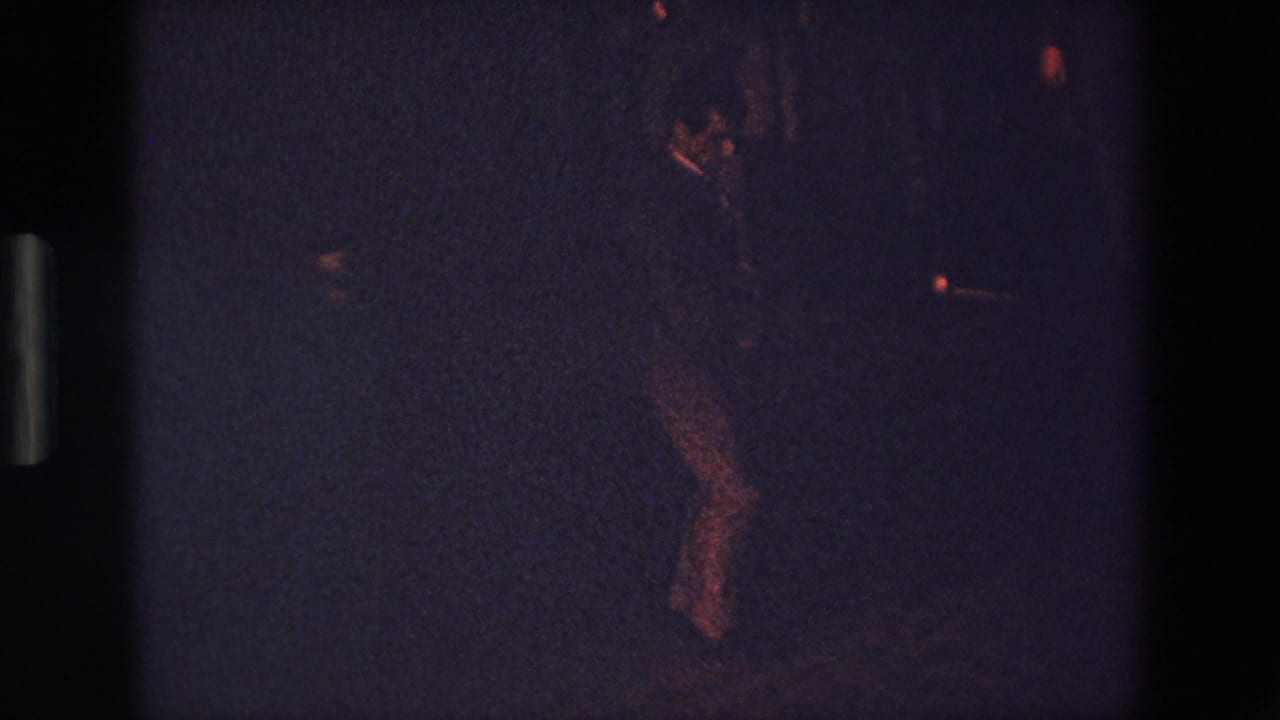Dim figure leaning or moving within a dark grainy frame.