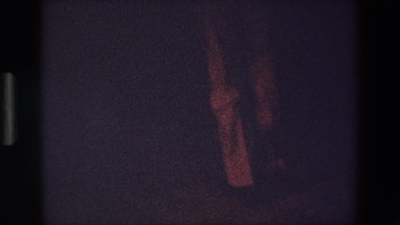In this Palermo 1967 8mm home-movie frame, a person's lower legs and shoes appear in reddish low light against a dark floor and background.