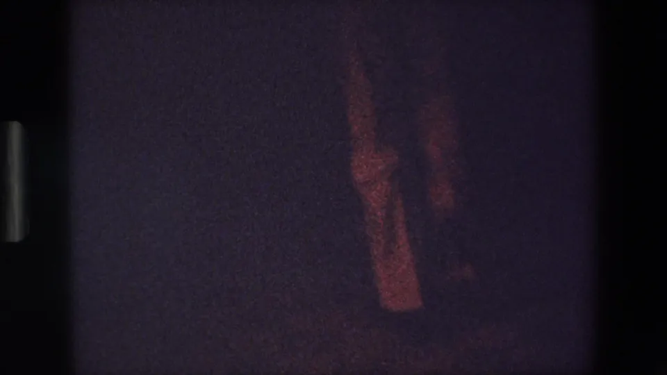 In this Palermo 1967 8mm home-movie frame, a person's lower legs and shoes appear in reddish low light against a dark floor and background.