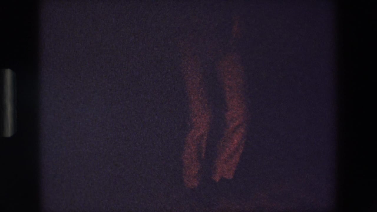 In this 1967 Palermo 8mm home-movie frame, a man's legs appear mid-step on a dim dance floor with heavy grain and faint reddish illumination.