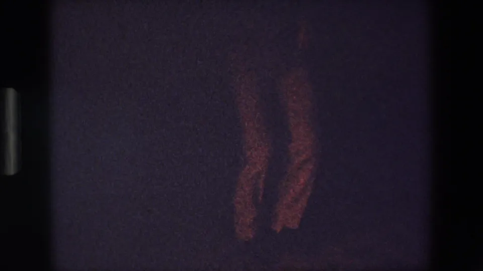In this 1967 Palermo 8mm home-movie frame, a man's legs appear mid-step on a dim dance floor with heavy grain and faint reddish illumination.
