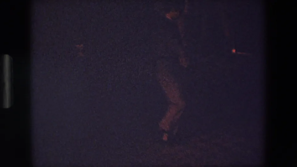 The 8mm frame shows the dancer's legs blurred by movement on a dark floor, with low exposure and faint red highlights consistent with indoor social lighting.