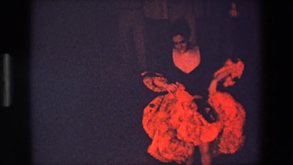 The performer turns sideways with arms and skirt in motion, the red costume standing out against the shadowed stage in the 8mm footage.
