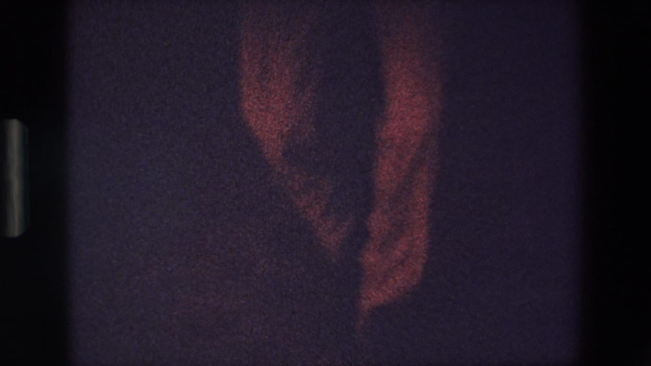 A dim red-lit 1967 Palermo home-movie frame shows part of a standing figure in a dark interior, with heavy grain and visible film-frame edges.