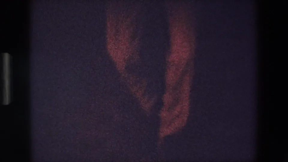 A dim red-lit 1967 Palermo home-movie frame shows part of a standing figure in a dark interior, with heavy grain and visible film-frame edges.