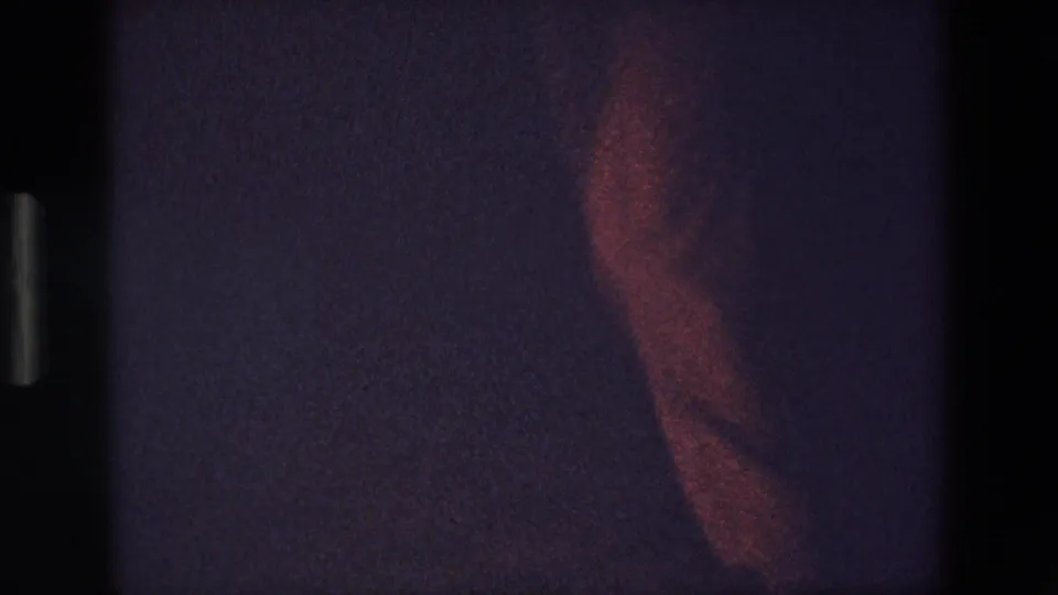 A partial figure appears under red-tinted light in a dark 8mm home-movie frame from the Palermo 1967 clip, with the person partly cropped by shadow and frame edge.