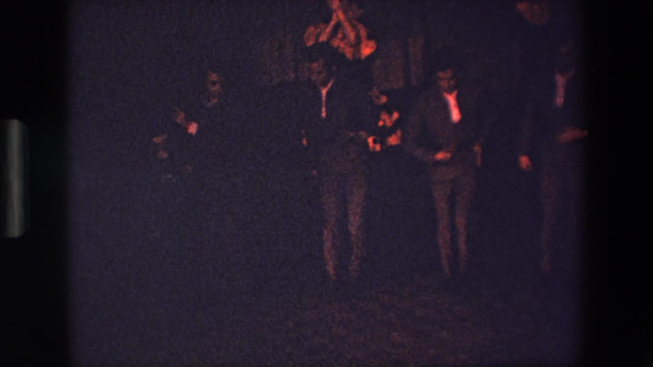Men in suits stand and move together in a dark performance space.