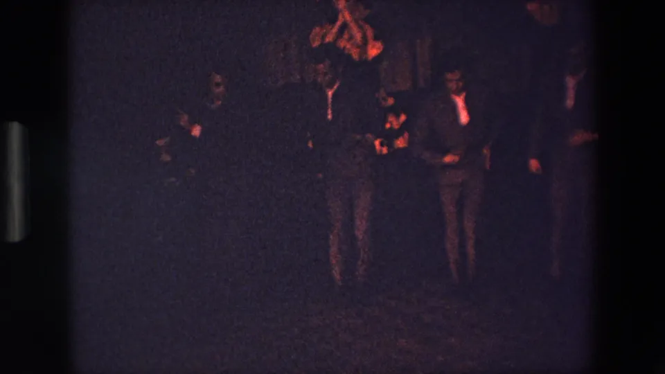 A small group of suited male performers or dancers is visible in the dim interior, with faces partly obscured by shadow and red-orange light.