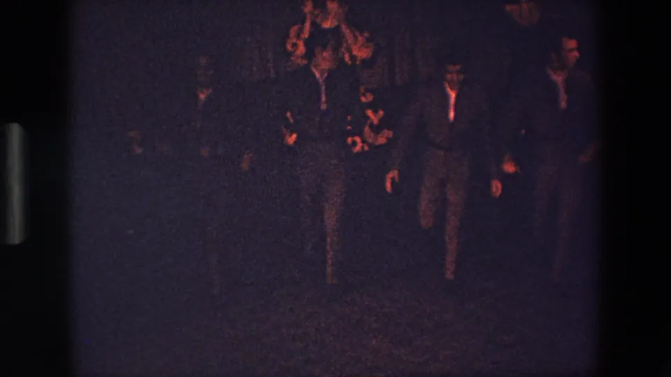A central suited figure appears to move forward or dance while other men in suits remain nearby in the dark, warm-lit club setting.