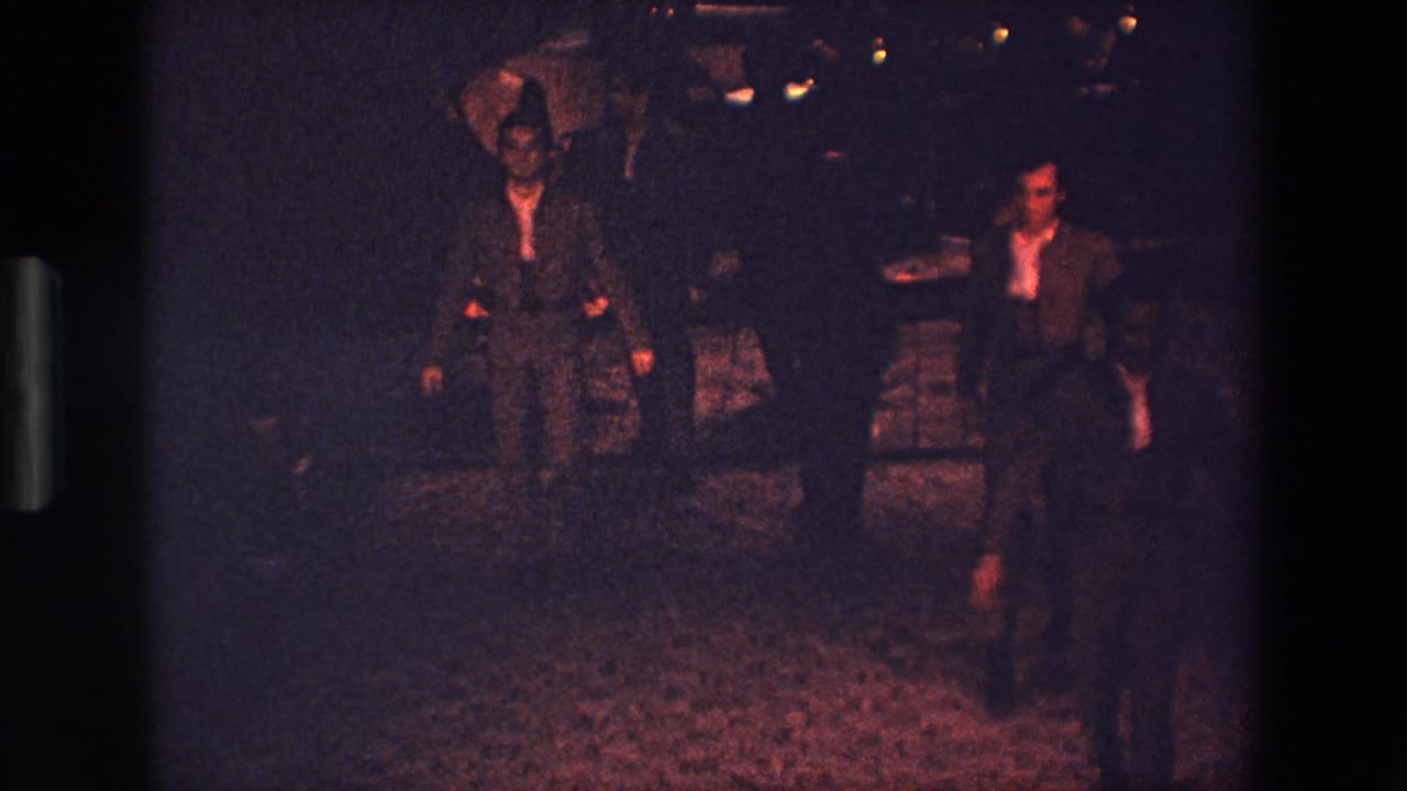 Two men in dark suits and white shirts move through a nighttime outdoor dance area in the Palermo 1967 home-movie footage, with faint lights and shadowed figures behind them.
