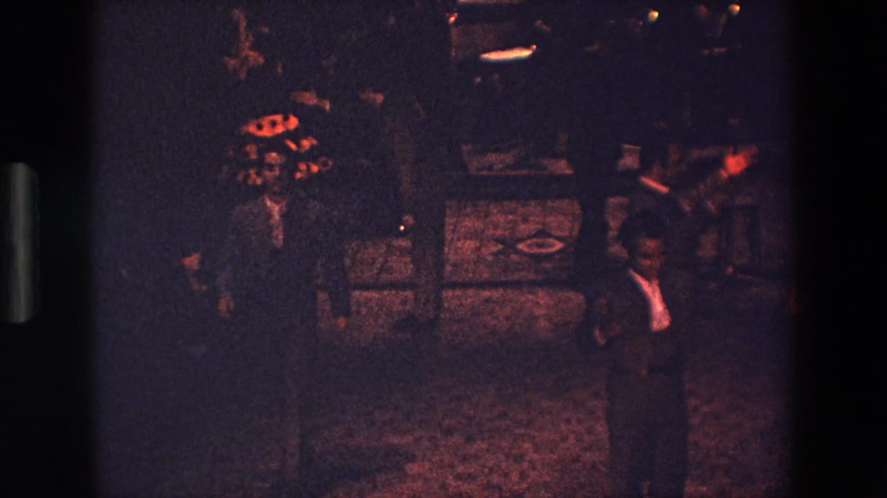 A man in a dark suit and white shirt stands near the foreground during the nighttime Palermo dance scene, with additional figures and ground markings partly visible in the dim frame.
