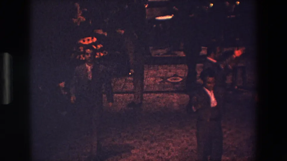A man in a dark suit and white shirt stands near the foreground during the nighttime Palermo dance scene, with additional figures and ground markings partly visible in the dim frame.