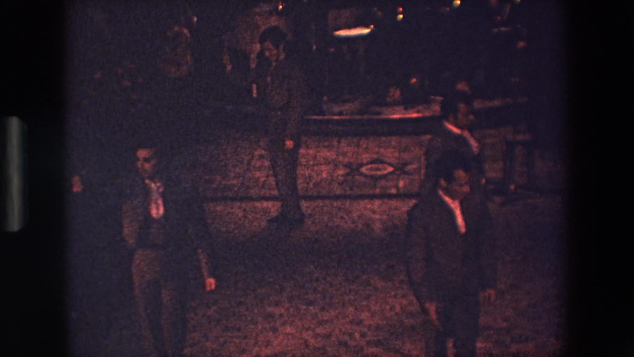 In 1967 Palermo 8mm footage, suited men stand in a low-lit interior with a patterned floor and a dark furnishing behind them.