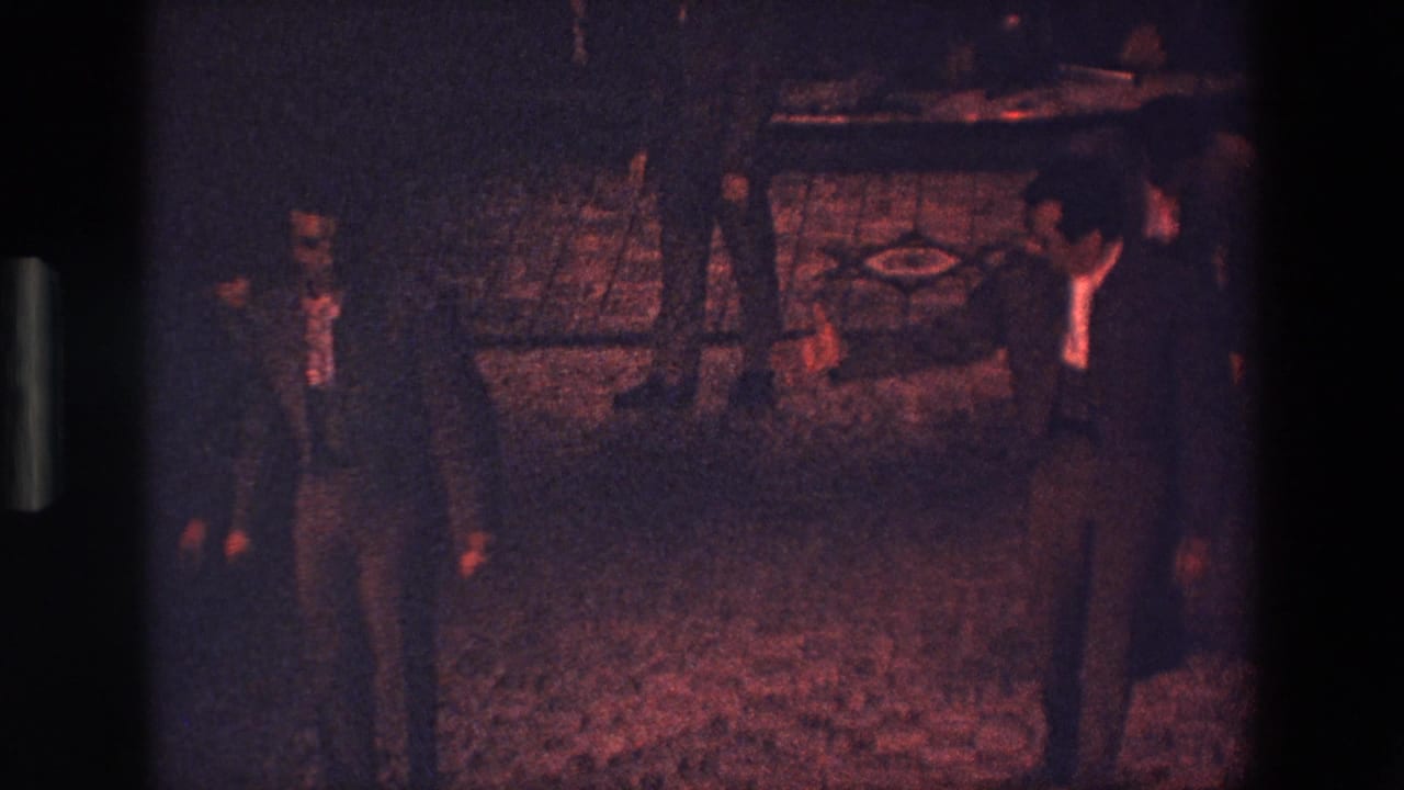 Several men in formal suits appear spaced across a dim Palermo interior, with low reddish light and a patterned floor visible in the 8mm frame.