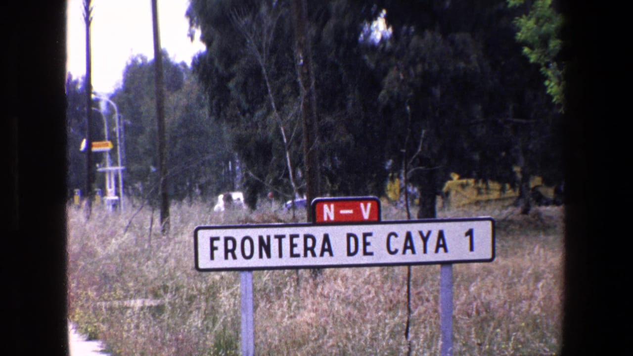 1967 Palermo-area 8mm frame showing a white roadside sign reading 'FRONTERA DE CAYA 1,' a red 'N-V' marker, dry grass, trees, utility poles, and a distant vehicle.