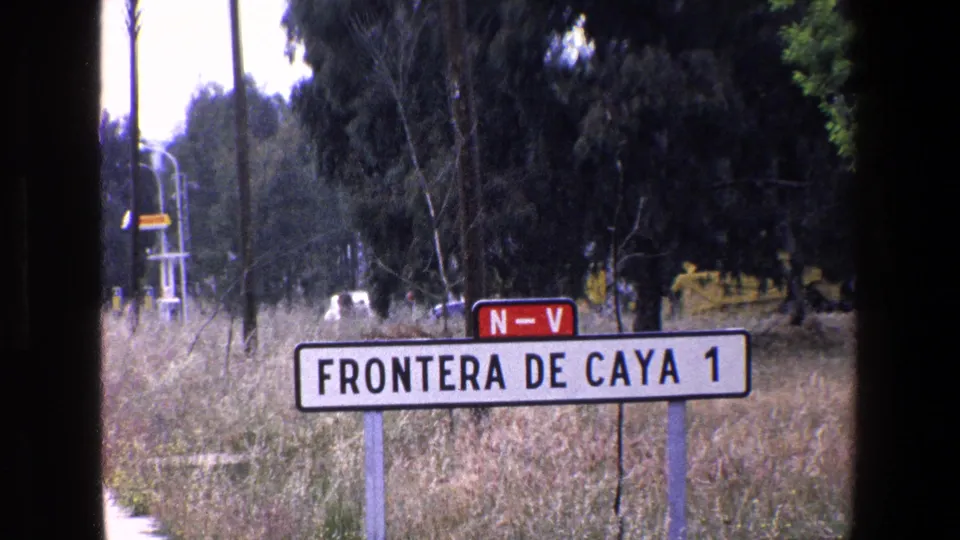 1967 Palermo-area 8mm frame showing a white roadside sign reading 'FRONTERA DE CAYA 1,' a red 'N-V' marker, dry grass, trees, utility poles, and a distant vehicle.