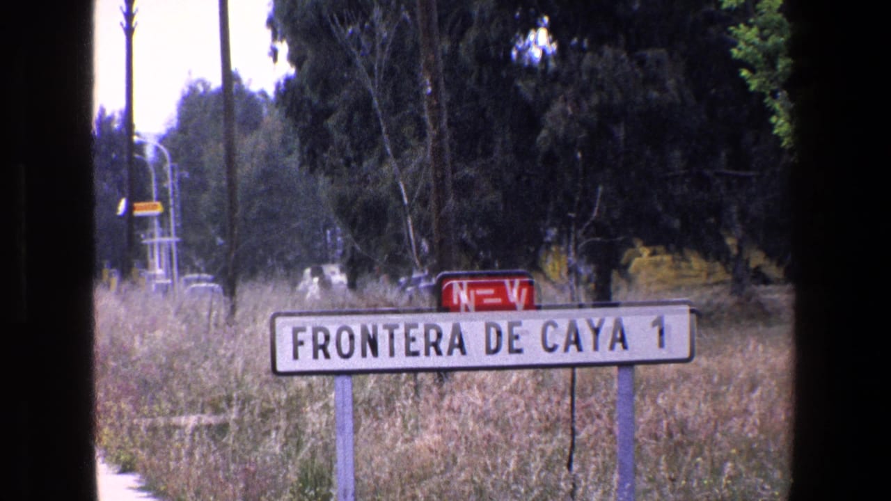 1967 Palermo-area 8mm frame showing the 'FRONTERA DE CAYA 1' sign near the bottom of the frame, with the red marker, dry vegetation, a pale road edge, trees, and utility poles behind it.