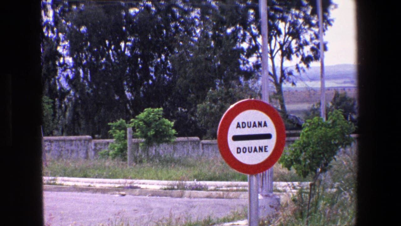1967 8mm frame showing a circular red-and-white customs sign reading ADUANA DOUANE near pavement, roadside plants, trees, and distant open land.