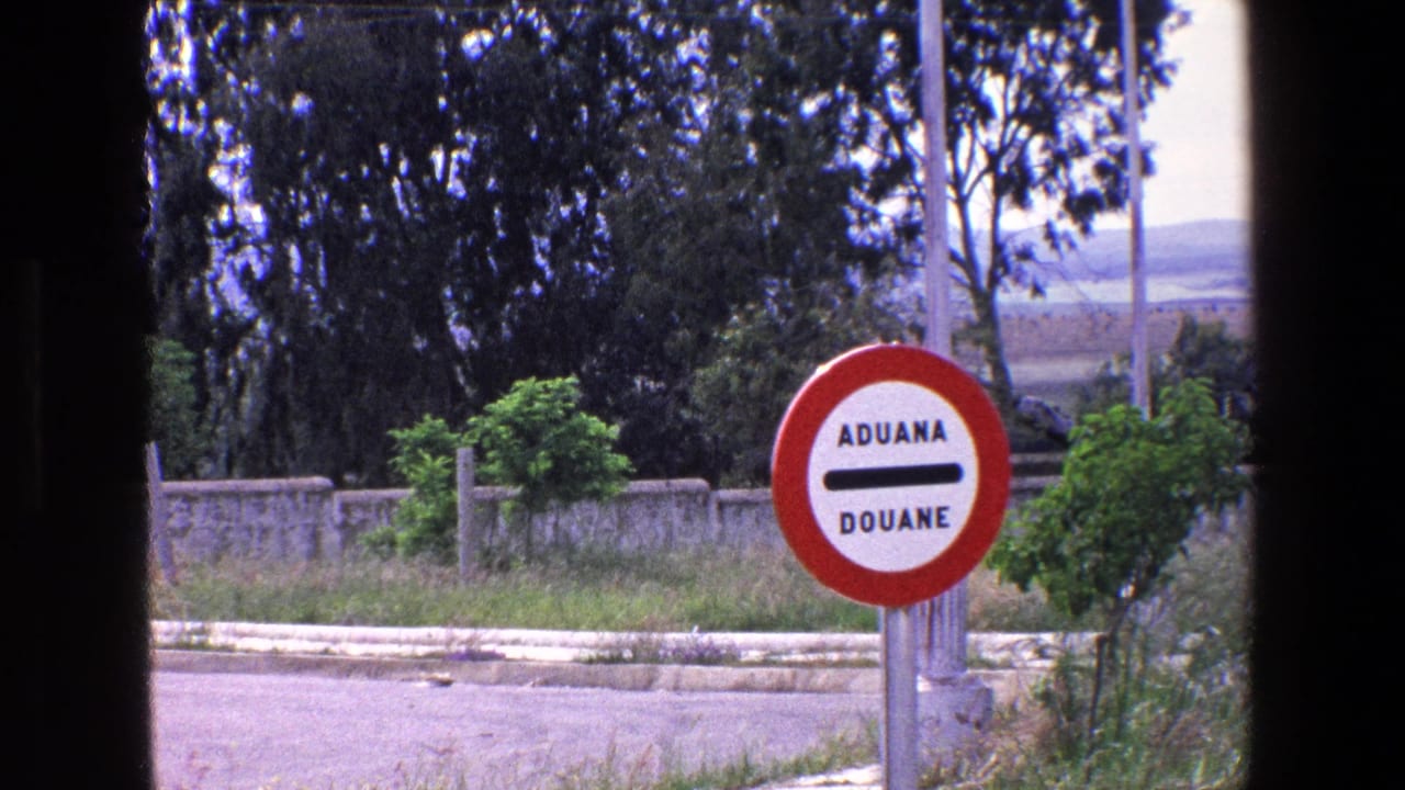 1967 Palermo-area frame with the ADUANA DOUANE sign centered near vegetation, a low wall, and open landscape beyond the road.