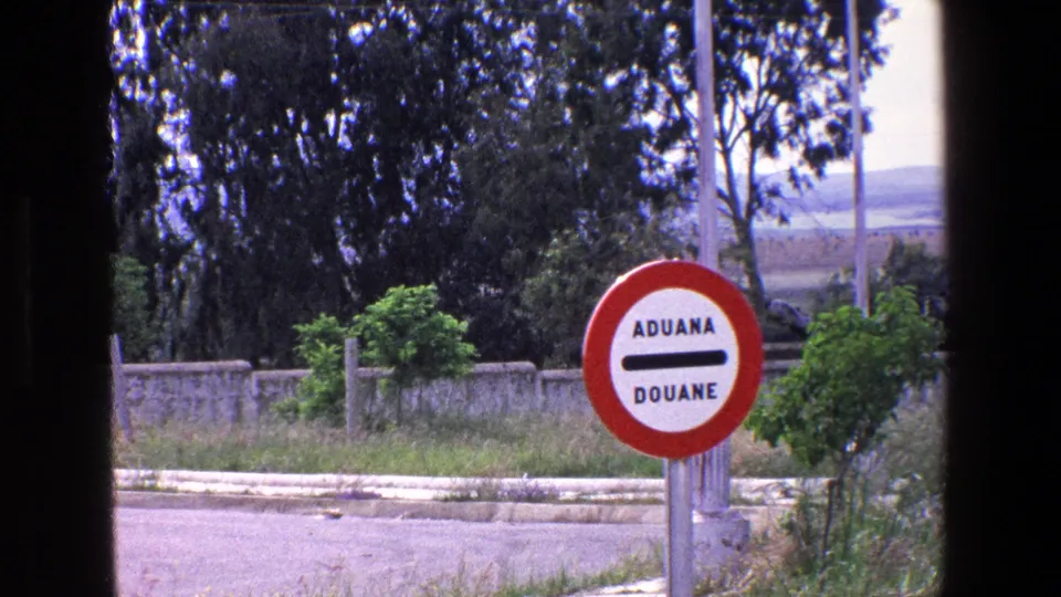 1967 Palermo-area frame with the ADUANA DOUANE sign centered near vegetation, a low wall, and open landscape beyond the road.