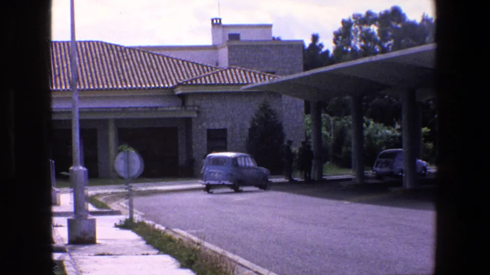 1967 Palermo-area frame showing a small vintage car on the paved approach beside a checkpoint canopy, tiled-roof building, trees, and distant pedestrians.