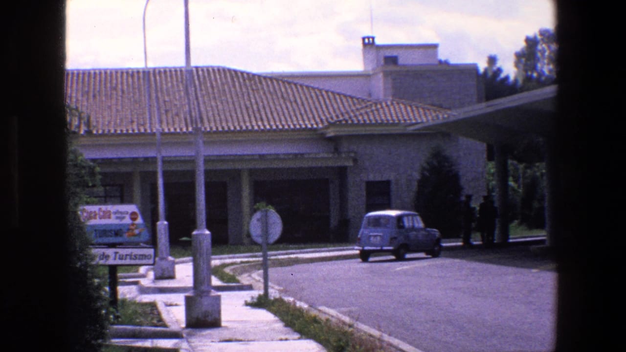 1967 Palermo home-movie frame showing a blue compact car on a roadway outside a tiled-roof building, with street signs, lamps, shrubs, and a covered parking area partly framed by dark edges.