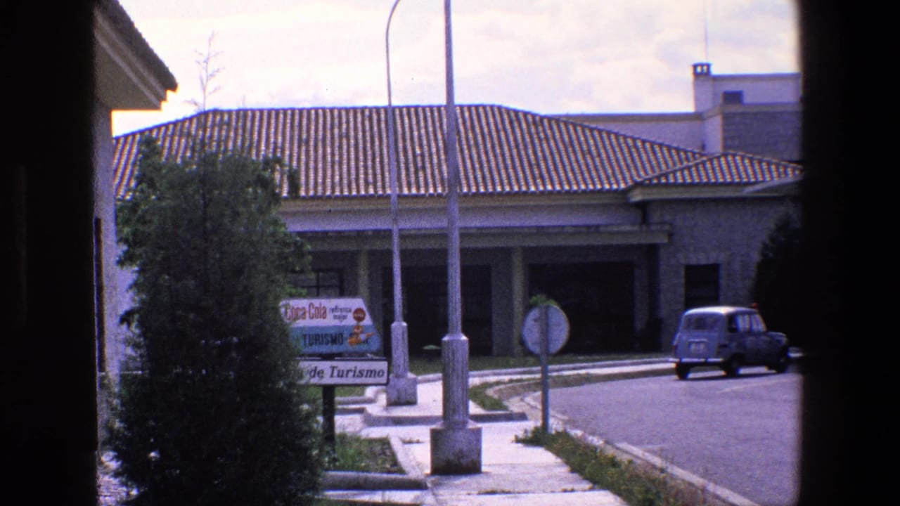 Roadside building with tourism signage, street lamps, shrubs, and a small blue car.