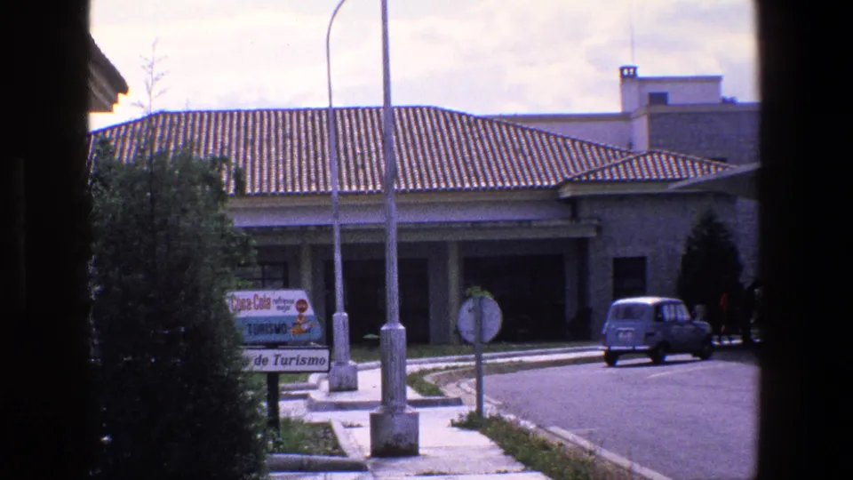 1967 Palermo street view showing the blue compact car farther along the road, a tiled-roof building, Turismo and Coca-Cola signage, a round road sign, and a dark vertical obstruction from the camera position.