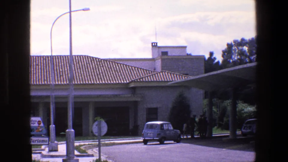 1967 Palermo frame showing a small blue car near a covered parking canopy where several people stand in shadow, with a tiled-roof building, roadside signs, street lamps, and dark window-like borders.