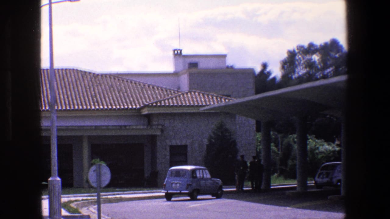 1967 Palermo street view with a small blue car near a white stone building, tiled roof, covered entrance, trees, and dark foreground framing.