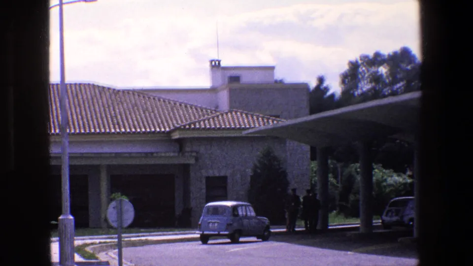 1967 Palermo street view with a small blue car near a white stone building, tiled roof, covered entrance, trees, and dark foreground framing.