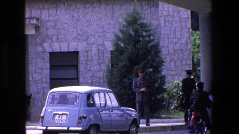 1967 Palermo street frame with a small blue car, two uniformed figures, a motorcyclist at the curb, stone building wall, window, and greenery.