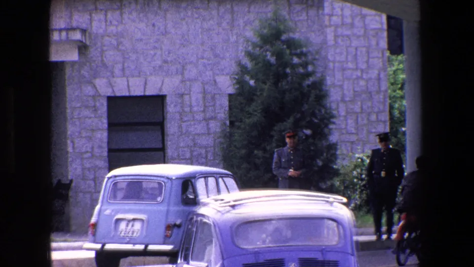 1967 Palermo exterior with a passing light-colored car in the foreground, a parked blue car, uniformed soldiers, stone facade, trees, and a motorcyclist at the right edge.