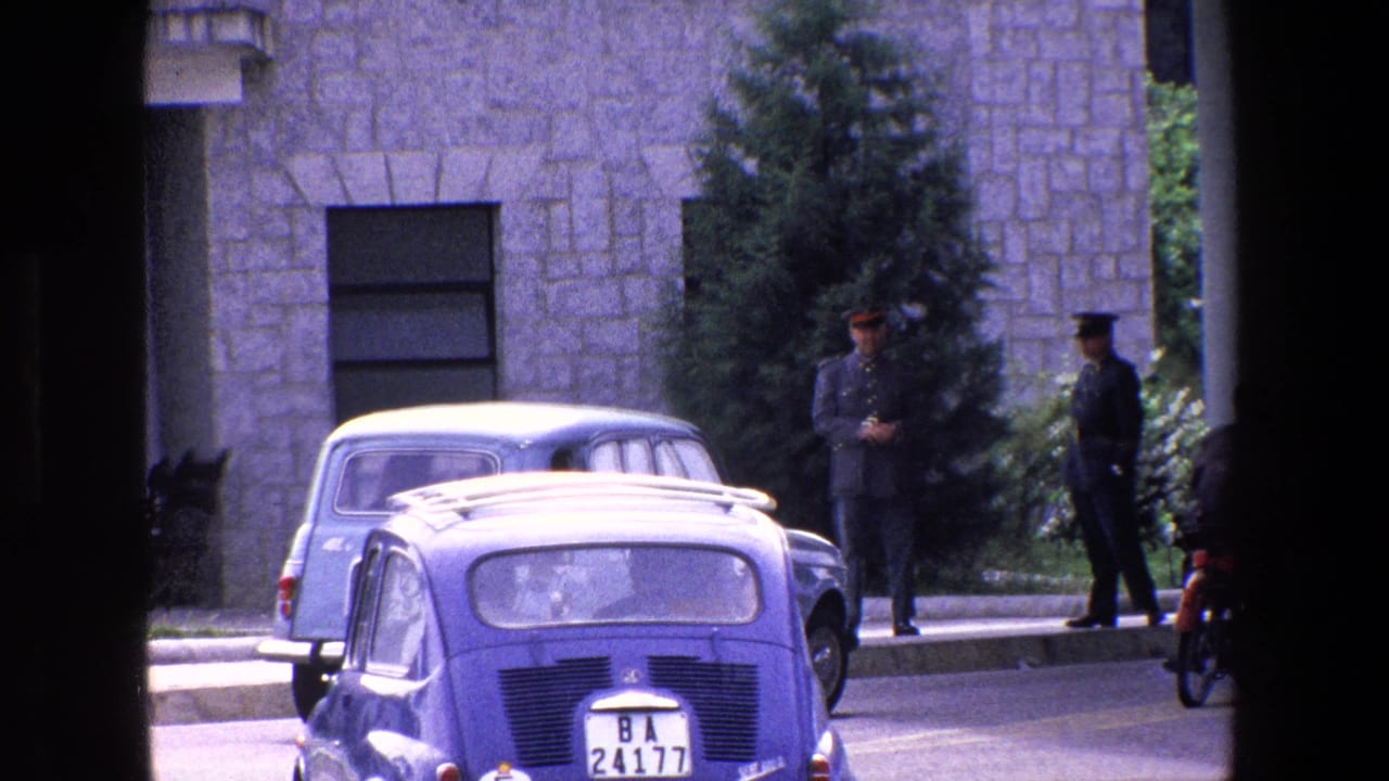 1967 Palermo street view with small parked European cars, two uniformed men on the sidewalk, and a stone building seen through dark foreground framing.