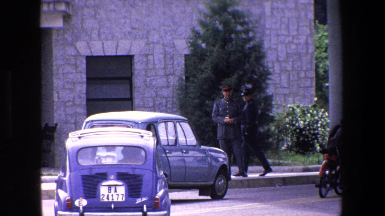 Small blue car in the street with uniformed men near a stone facade.