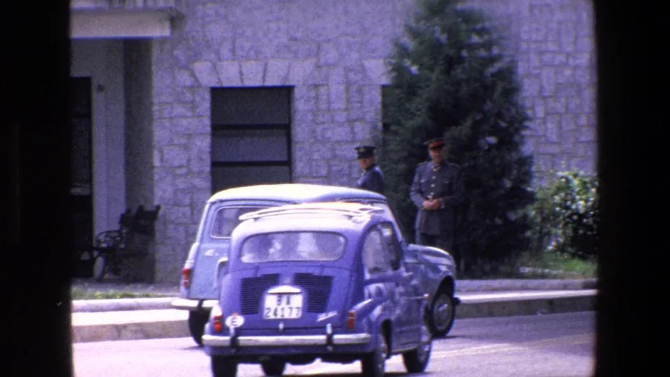 1967 Palermo footage showing the rear of a small blue car in the street, a parked light blue car, and uniformed men near the curb.