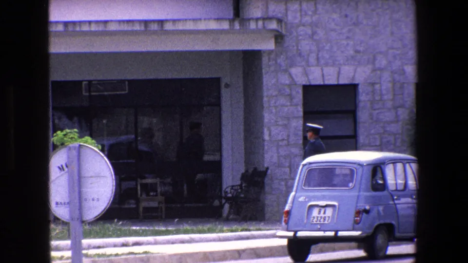1967 Palermo street view with a compact car near a shaded building entrance, a road sign in the foreground, and a uniformed man standing by the curb.