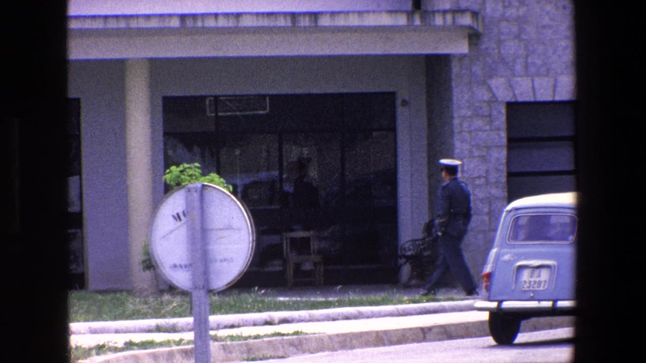 1967 Palermo home-movie frame showing a uniformed man in a peaked cap walking near glass doors of a stone-fronted building, partly framed by dark window edges.