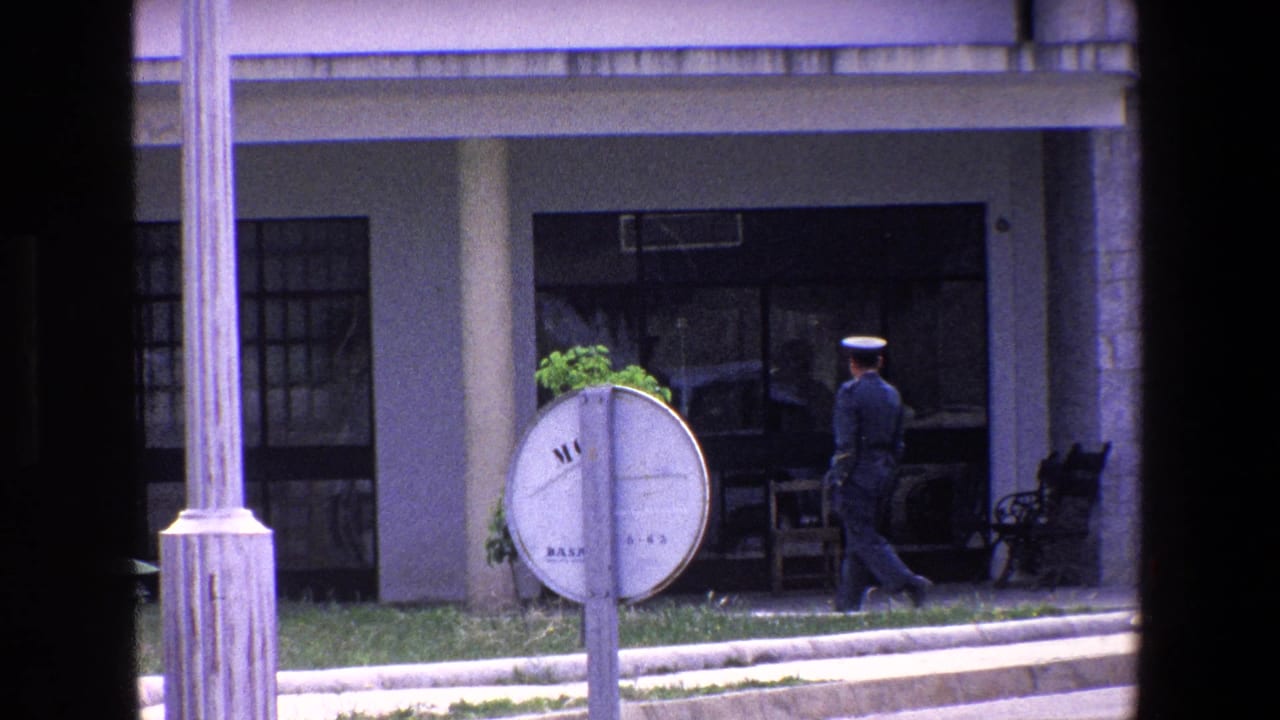 1967 Palermo frame with a uniformed official moving leftward along a stone and glass building front, seen through a dark vertical window frame.