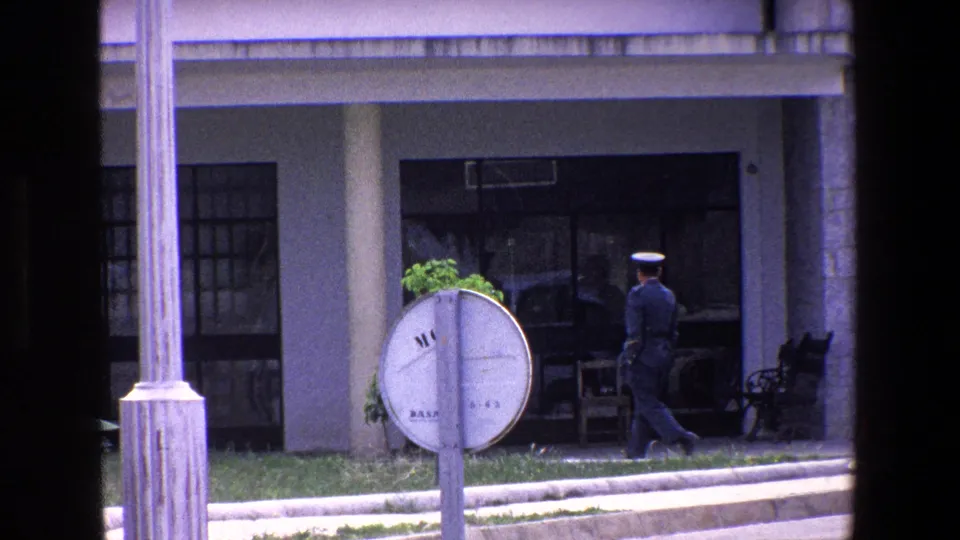 1967 Palermo frame with a uniformed official moving leftward along a stone and glass building front, seen through a dark vertical window frame.