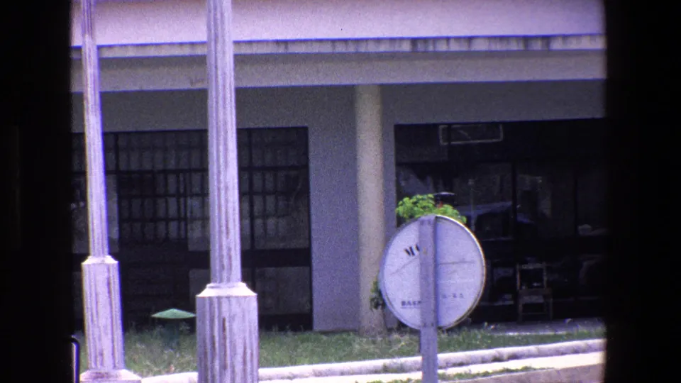 1967 Palermo home-movie frame focused on the building entrance, circular roadside sign, stone columns, and dark window framing after the uniformed figure has moved out of the main view.