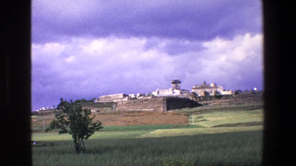 1967 Palermo-area travel frame showing a distant hilltop settlement or complex above green fields beneath a dramatic cloudy sky, with a small tree in the foreground.