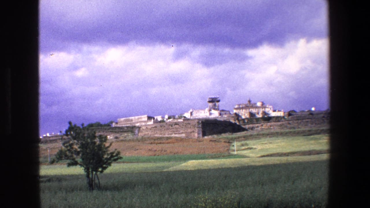 1967 8mm home-movie frame from Palermo, Italy, showing a distant historic stone settlement with pale buildings and a tower beyond fields and a lone tree.