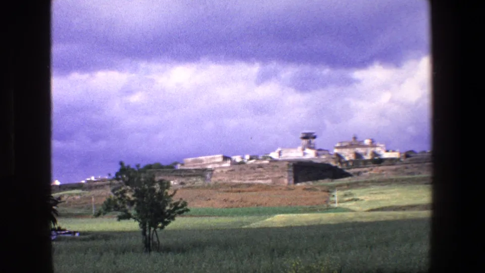 1967 Palermo travel footage frame showing a distant stone-walled complex or settlement across open countryside, filmed through a dark window-like frame.