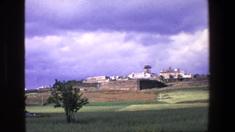 1967 8mm frame from Palermo, Italy, with a lone tree and fields leading to distant stone walls, white structures, and a tower under a cloud-heavy sky.