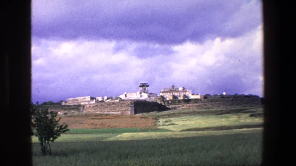 Final 1967 Palermo 8mm frame showing the distant historic stone settlement, tower, green fields, and heavy clouds, with the scene partially bordered by dark window framing.