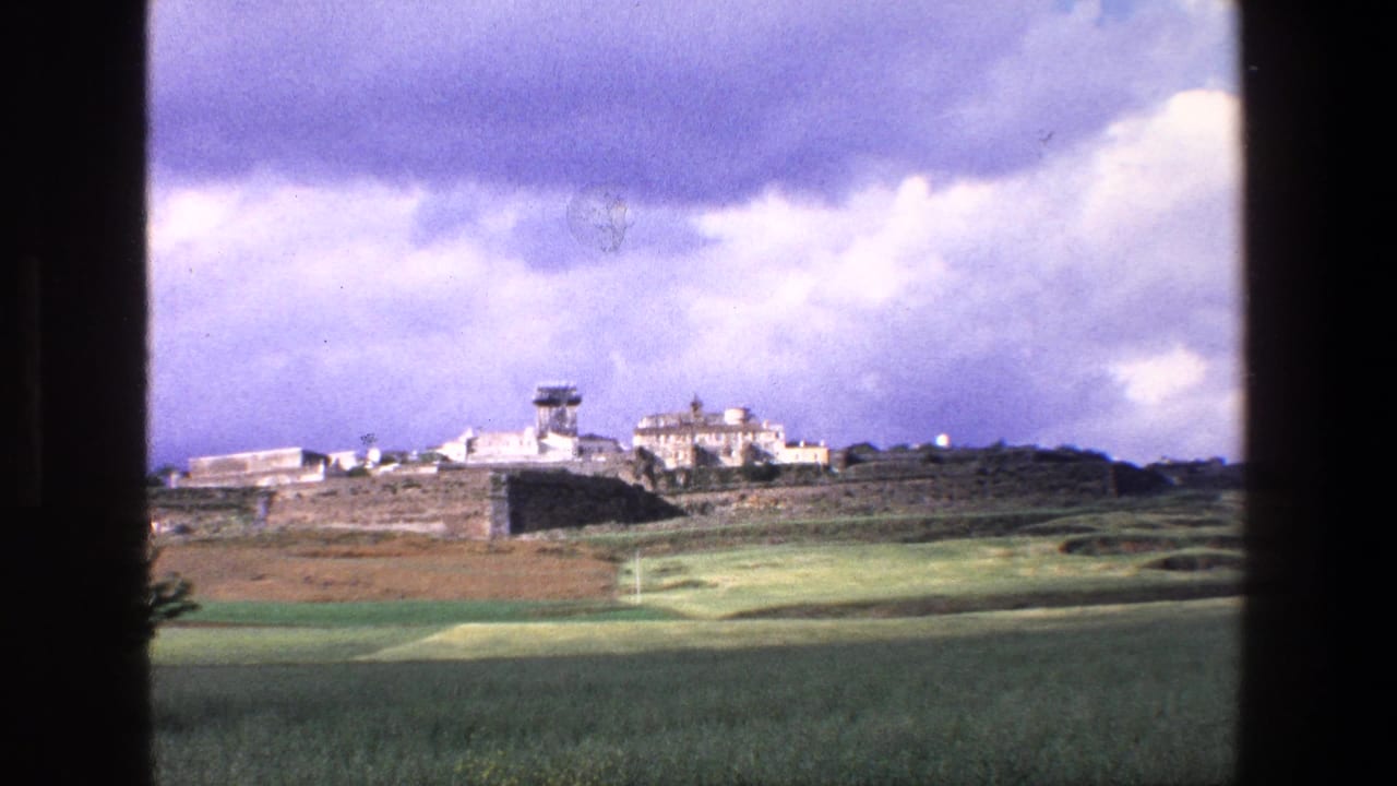 1967 Palermo-area countryside view through a window frame, with rolling fields, pale hilltop buildings, and a tower beneath a cloudy purple-tinged sky.