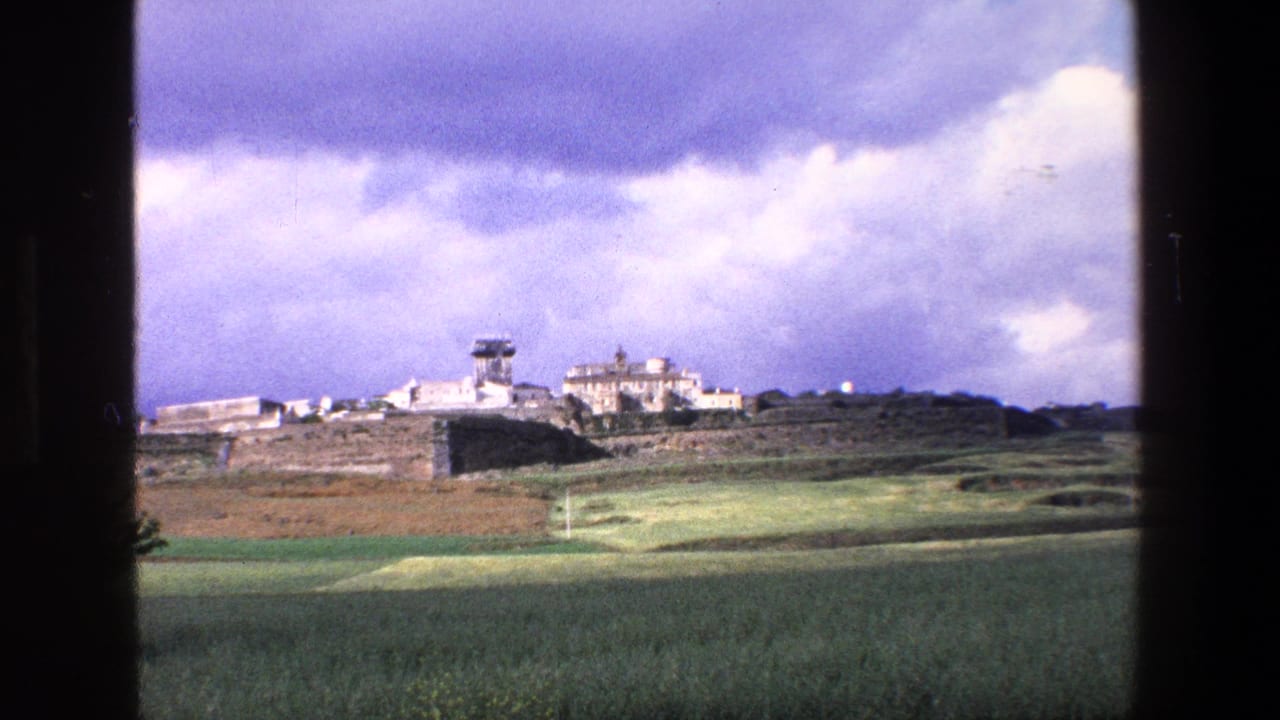 1967 Palermo-area rural landscape framed by a dark window mullion, showing fields, a low embankment, pale buildings, and a tower under a dramatic sky.