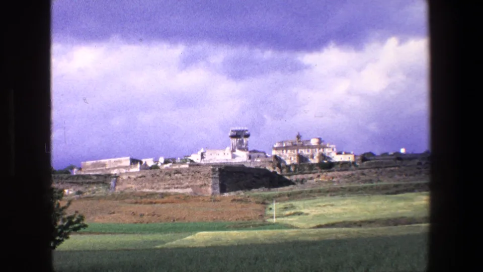 1967 Palermo-area home-movie frame showing a closer hillside view of pale buildings, a tower, open fields, and a dark window edge in the foreground.
