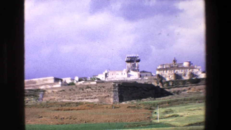 1967 Palermo-area countryside frame with the camera shifted right, showing pale hilltop buildings, a prominent tower, fields, and the edge of a dark window frame.
